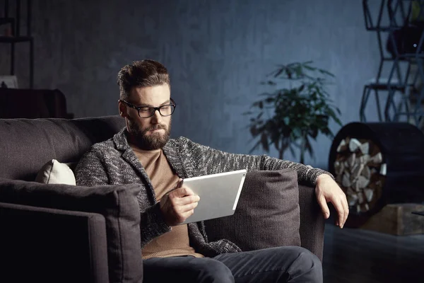 Handsome attractive elegant classic man wearing trendy grey and glasses, with beard, sitting on luxury furniture in fashionable dark interior with laptop