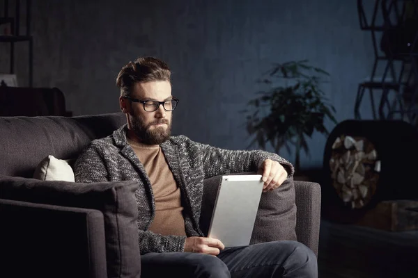 Handsome attractive elegant classic man wearing trendy grey and glasses, with beard, sitting on luxury furniture in fashionable dark interior with laptop