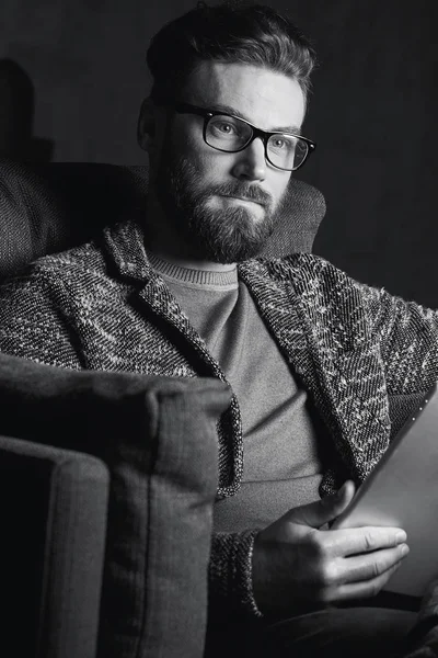 Porträtt av stiliga attraktiva eleganta klassiska man bär trendiga grå och glasögon, med skägg, sitter på lyxiga möbler i fashionabla mörka interiören med laptop i svart och vitt — Stockfoto