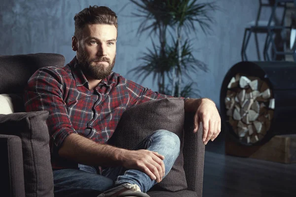 Handsome attractive elegant classic man wearing trendy shirt, with beard, sitting and having rest on luxury furniture in fashionable dark interior