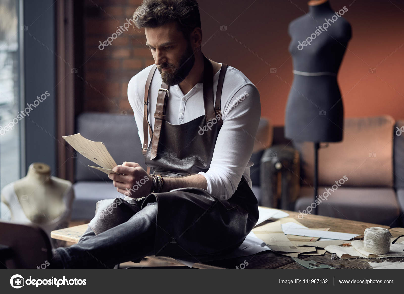 Young handsome tailor with beard in white shirt and leather apron sitting on wooden table with threads in amazing atelier with antique furniture and mannequin on background Stock Photo mochak 14198713...
