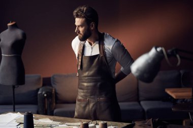 Antika mobilyalar ve manken arka plan üzerinde şaşırtıcı atelier konular ile beyaz gömlek ve deri önlük ayakta ahşap masa yakınındaki sakallı genç yakışıklı terzi