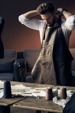 Antika mobilyalar ve manken arka plan üzerinde şaşırtıcı atelier konular ile beyaz gömlek ve deri önlük ayakta ahşap masa yakınındaki sakallı genç yakışıklı terzi