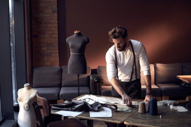 Sakal ve gözlük kahverengi deri jartiyer konu, önlük ve antika mobilyalar ve manken şaşırtıcı atelier makas ile ahşap masa yakınındaki arka plan üzerinde çalışma ile Beyaz gömlekli güzel ciddi terzi