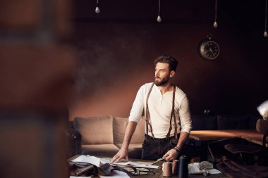 Sakal ve gözlük konu, önlük ve makas ile ahşap masa yanında duran ve antika mobilyalarla harika atelier penceresinde bakarak kahverengi deri pantolon askısı ile Beyaz gömlekli güzel ciddi terzi 