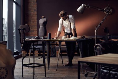 Kahverengi deri jartiyer konu, önlük ve antika mobilyalar ve manken şaşırtıcı atelier makas ile ahşap masa yakınındaki arka plan üzerinde çalışma ile Beyaz gömlekli sakallı yakışıklı ciddi terzi