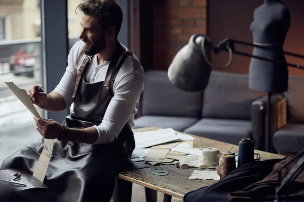 Joven sastre hermoso en camisa blanca y delantal de cuero sentado en mesa de madera con hilos y dibujo en atelier increíble — Foto de Stock