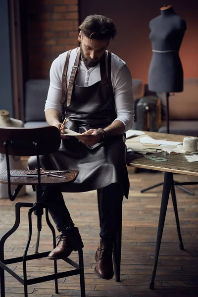 Young handsome tailor with beard in white shirt and leather apron sitting on wooden table with threads and drawing in amazing atelier with antique furniture — Stock Photo, Image