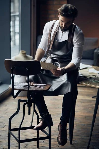 Young handsome tailor with beard in white shirt and leather apron sitting on wooden table with threads and drawing in amazing atelier with antique furniture
