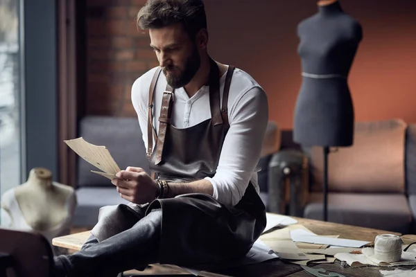 Joven sastre guapo con barba en camisa blanca y delantal de cuero sentado en mesa de madera con hilos en atelier increíble con muebles antiguos y maniquí en el fondo — Foto de Stock