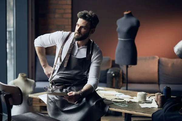 Young handsome tailor with beard in white shirt and leather apron sitting on wooden table with threads and thinking in amazing atelier with antique furniture and mannequin on background