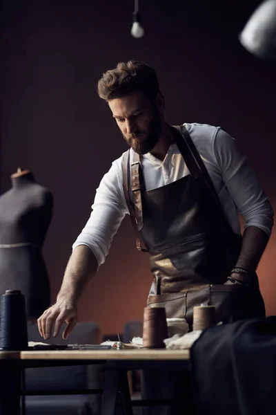 Fiatal jóképű török szakállas fehér inget és bőr kötény ollóval zsebében dolgozik közelében csodálatos műtermében, antik bútorokkal és manöken szálak fából készült asztal háttere — Stock Fotó