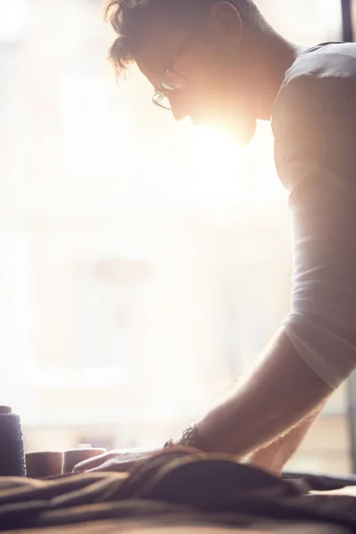 Joven sastre guapo con barba y gafas en camisa blanca trabajando cerca de mesa de madera con hilos en atelier increíble con muebles antiguos y gran luz del sol — Foto de Stock