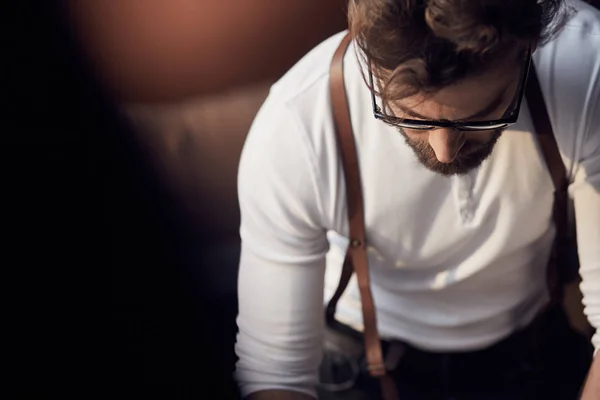 Joven sastre serio con barba y gafas en camisa blanca con tirantes de cuero trabajando cerca de mesa de madera con hilos en increíble atelier con muebles antiguos — Foto de Stock