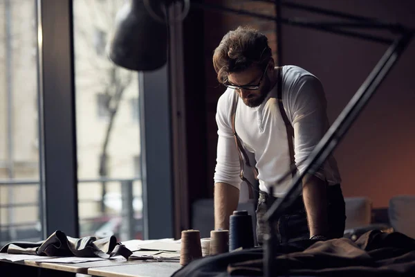 Young serious tailor with beard and glasses in white shirt with leather suspenders looking on drawing near wooden table with threads in amazing atelier with antique furniture and mannequin on background — Stock Photo, Image