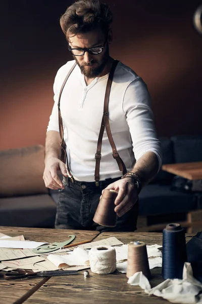 Young cute tailor with beard and glasses in white shirt with leather suspenders working near wooden table with threads in amazing atelier with antique furniture and mannequin on background — Stock Photo, Image