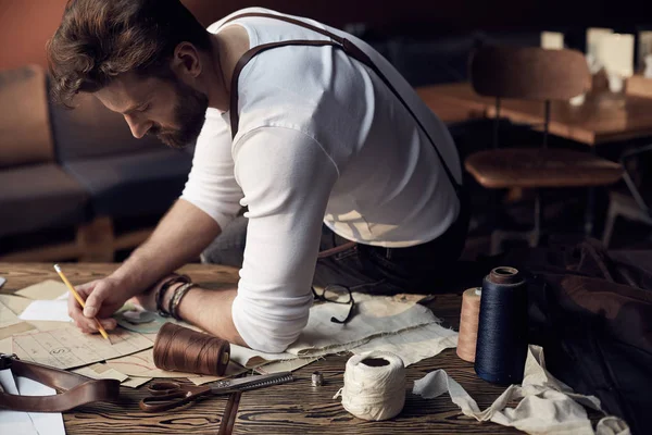 Guapo sastre hombre con barba en camisa blanca con tirantes de cuero marrón trabajando cerca de mesa de madera con hilos en atelier increíble con muebles antiguos y maniquí en el fondo — Foto de Stock