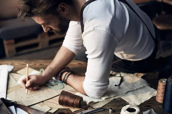 Hermoso sastre hombre con barba en camisa blanca con tirantes de cuero marrón dibujo cerca de la mesa de madera con hilos y tijeras en atelier increíble con muebles antiguos y maniquí en el fondo — Foto de Stock