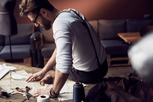 Handsome tailor male with beard and glasses in white shirt with brown leather suspenders drawing near wooden table with threads, apron and scissors in amazing atelier with antique furniture and mannequin on background