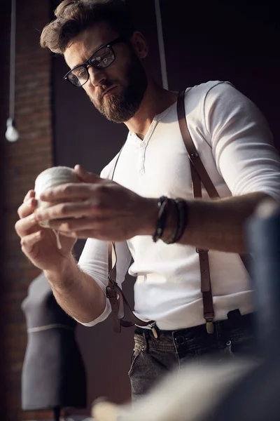 Guapo sastre hombre con barba y gafas en camisa blanca con tirantes de cuero marrón trabajando en increíble atelier con muebles antiguos y maniquí sobre fondo — Foto de Stock