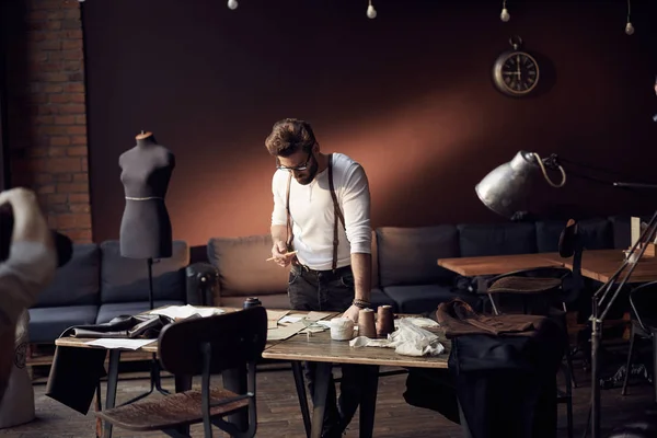 Guapo sastre hombre con barba y gafas en camisa blanca con tirantes de cuero marrón trabajando cerca de mesa de madera con hilos, delantal y tijeras en increíble atelier con muebles antiguos y maniquí sobre fondo — Foto de Stock