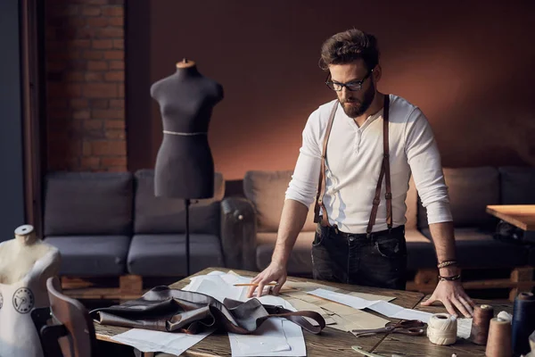 Lindo sastre masculino con barba y gafas en camisa blanca con tirantes de cuero marrón trabajando cerca de mesa de madera con hilos, delantal y tijeras en increíble atelier con muebles antiguos y maniquí en el fondo —  Fotos de Stock