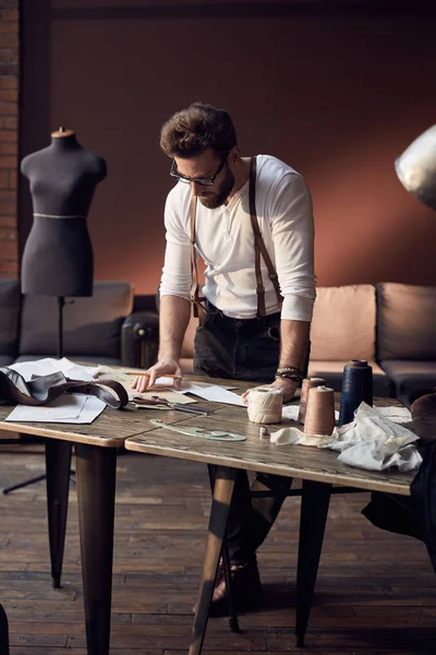Aranyos török férfi szakáll és poharak a fehér ing, barna bőr harisnyatartó dolgozik, közel fából készült asztal, szálat, kötény és olló csodálatos műtermében, antik bútorokkal és a manöken a háttérben — Stock Fotó