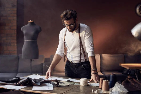 Handsome smiling tailor with beard and glasses in white shirt with brown leather suspenders working near wooden table with threads, apron and scissors in amazing atelier with antique furniture and mannequin on background — Stock Photo, Image