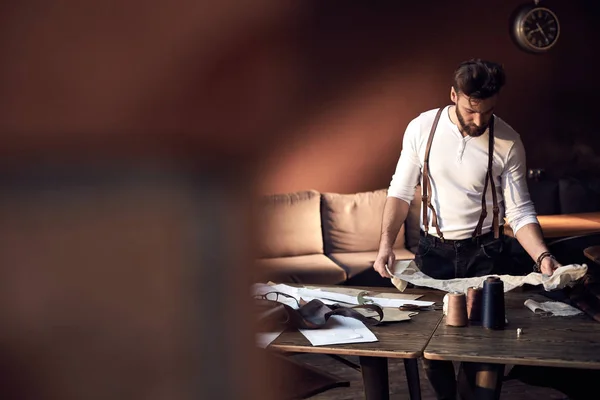 Beautiful serious tailor with beard and glasses in white shirt with brown leather suspenders working near wooden table with threads, apron and scissors in amazing atelier with antique furniture — Stock Photo, Image