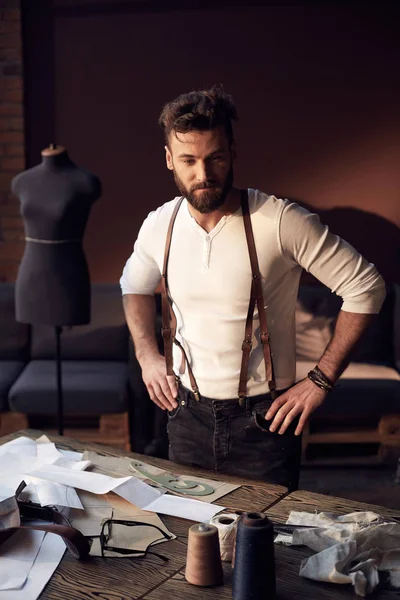 Beautiful serious tailor with beard in white shirt with brown leather suspenders standing near wooden table with threads, apron and scissors and thinking in amazing atelier with antique furniture — Stock Photo, Image