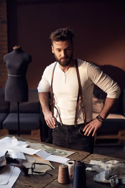 Bellissimo sarto serio con barba in camicia bianca con bretelle in pelle marrone in piedi vicino al tavolo in legno con fili, grembiule e forbici e pensando in incredibile atelier con mobili antichi — Foto Stock