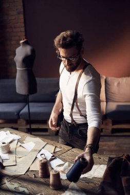 Sakal ve gözlük ahşap masa konu ile yakın çalışma ve antika mobilyalar ve manken şaşırtıcı atelier ekipman arka plan üzerinde dikiş kahverengi deri pantolon askısı ile beyaz gömlek büyük ciddi terzi