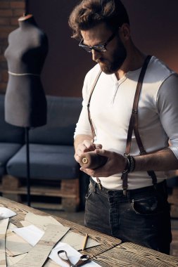 Sakal ve gözlük ahşap masa konu ile yakın çalışma ve antika mobilyalar ve manken şaşırtıcı atelier ekipman arka plan üzerinde dikiş kahverengi deri pantolon askısı ile beyaz gömlek ile şirin ciddi terzi