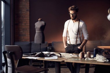 Sakal ve gözlük ahşap masa konu ile yakın duran ve antika mobilyalar ve manken şaşırtıcı atelier ekipman arka plan üzerinde terzilik kahverengi deri pantolon askısı ile Beyaz gömlekli ciddi terzi
