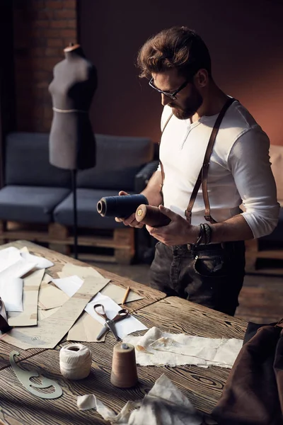 Great serious tailor with beard and glasses in white shirt with brown leather suspenders working near wooden table with threads and scissors in amazing atelier with antique furniture and mannequin on background — Stock Photo, Image