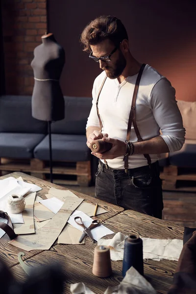 Great serious tailor with beard and glasses in white shirt with brown leather suspenders working near wooden table with threads and tailoring equipment in amazing atelier with antique furniture and mannequin on background — Stock Photo, Image