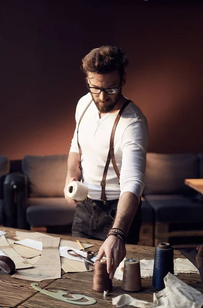 Serious tailor with beard and glasses in white shirt with brown leather suspenders working near wooden table with threads and tailoring equipment in amazing atelier with antique furniture — Stock Photo, Image