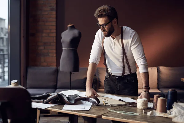 Amazing serious tailor with beard in white shirt with brown leather suspenders working near wooden table with threads, apron and scissors in great atelier with antique furniture and mannequin on background