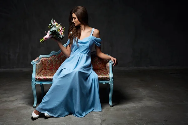 Cute young model female with dark hair in long blue dress sitting on fashionable sofa with flowers in studio — Stock Photo, Image