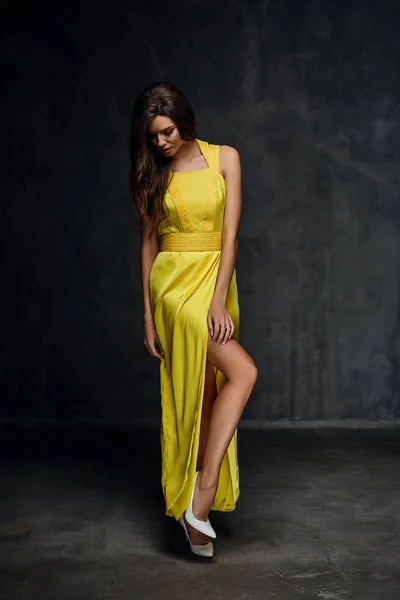 Ganska ung Sexig modell kvinna med mörkt hår i fantastisk lång gul klänning och vita skor poserar i mörka studio — Stockfoto