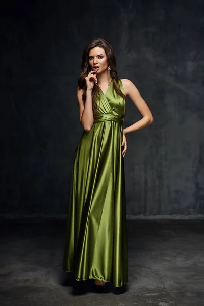 Pretty young sexy model female with dark hair in amazing long green glossy dress and white shoes posing in dark studio — Stock Photo, Image