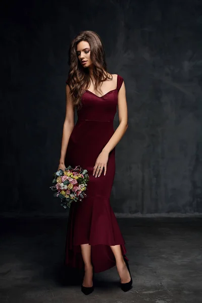 Ganska ung Sexig modell kvinna med mörkt hår i fantastiska långa cherry klänning och svarta skor poserar med blommor i mörka studio — Stockfoto