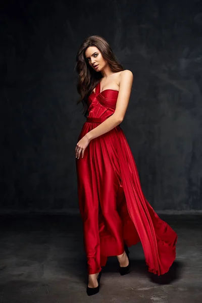 Pretty young sexy model female with dark hair in amazing long red dress and black shoes posing in dark studio — Stock Photo, Image