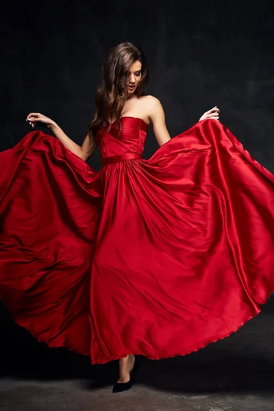 Pretty young sexy model female with dark hair in amazing long red draped dress and black shoes posing in dark studio — Stock Photo, Image