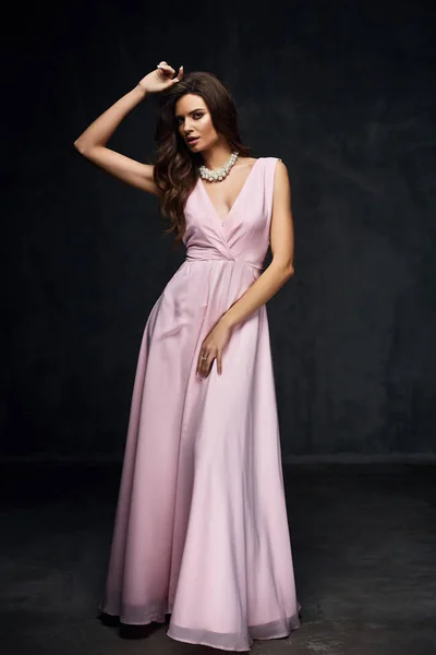 暗いスタジオでポーズをとって素晴らしい長い光のピンクのドレスに黒い髪を持つ美しい若いセクシーなモデル女性 — ストック写真