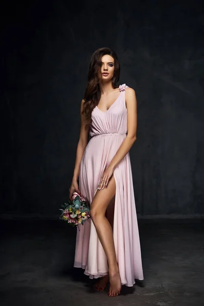 Beautiful young sexy model female with dark hair in gorgeous romantic long light pink dress posing with flowers in dark studio — Stock Photo, Image