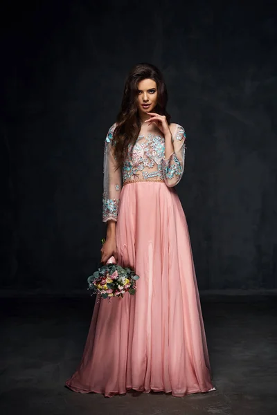 Beautiful young sexy model female with dark hair in gorgeous romantic glossy long light pink dress posing with flowers in dark studio — Stock Photo, Image