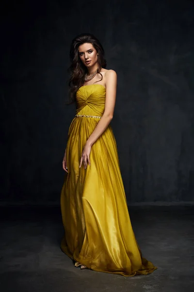 Szép fiatal szexi modell Sötét haja gyönyörű romantikus fényes hosszú világos arany női ruha, pózol, sötét stúdióban — Stock Fotó