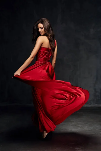 Досить молода сексуальна модель жінка з темним волоссям у дивовижній довгій червоній сукні та чорному взутті позує в темній студії Стокова Картинка