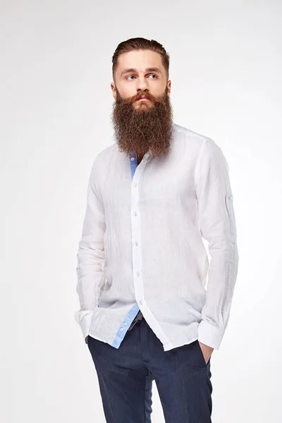 Молодий бородатий чоловік позує в студії в модній лляній білій сорочці і синіх штанях — стокове фото